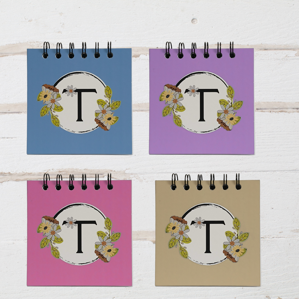 Four small spiral-bound notebooks with floral designs and letter 'T' on a white wooden surface.