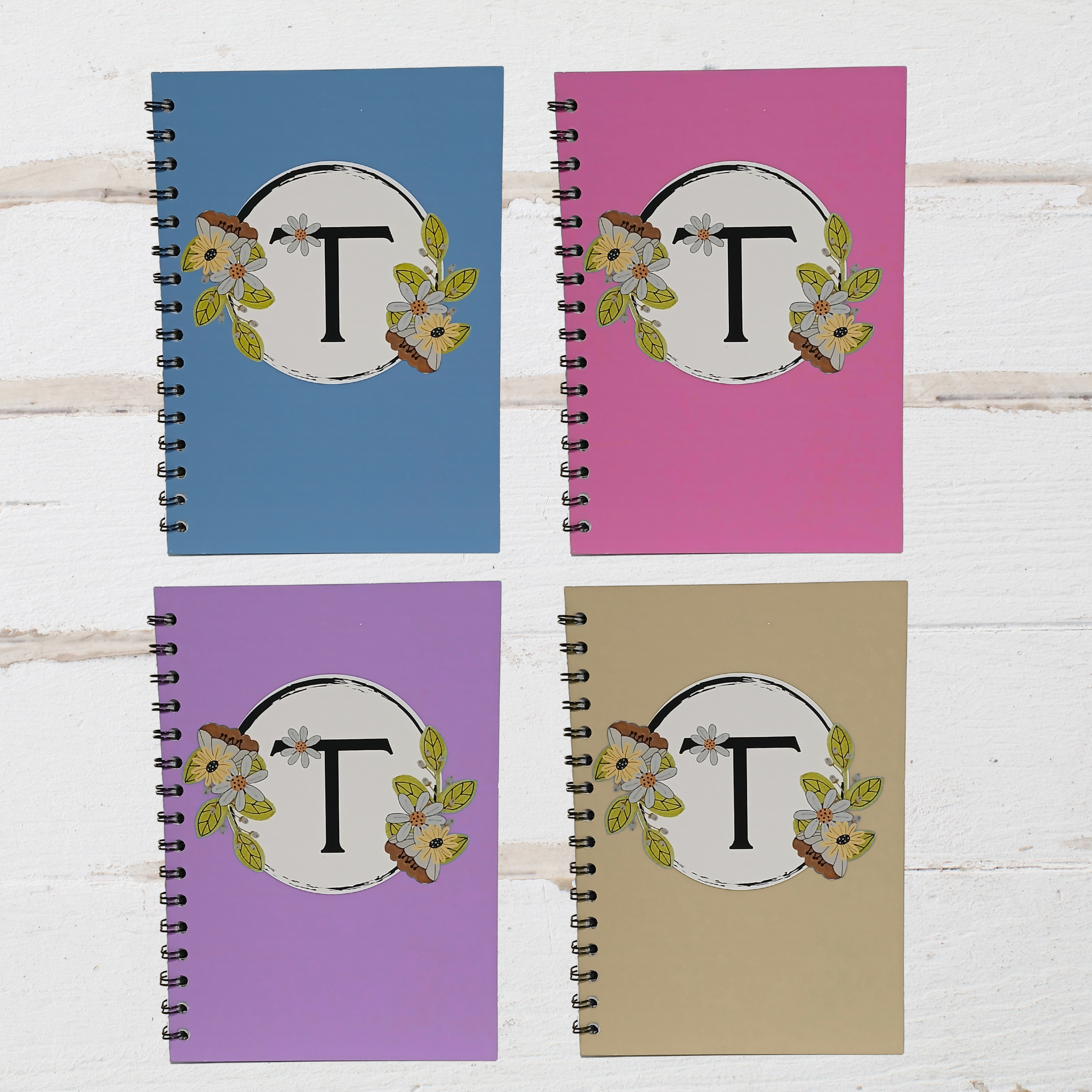 Four spiral-bound notebooks with floral designs and letter 'T' on a wooden surface.